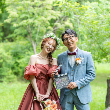 花冠をつけたテラコッタドレスの花嫁とライトブルースーツの新郎が、カチンコを持って並ぶ新緑の前撮り