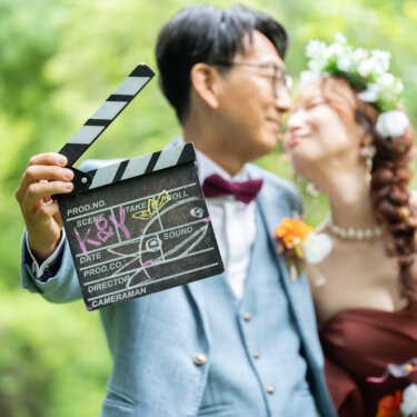 新緑を背に、ふたりの名前を書いたカチンコを手に顔を寄せ合う新郎新婦。映画のワンシーン風のフォトウェディング