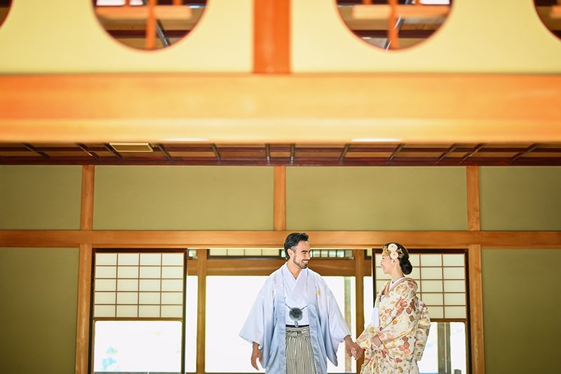 円窓のある伝統建築の広間で手をつなぎ見つめ合う、水色の紋付羽織の新郎と生花を髪に飾った着物姿の花嫁