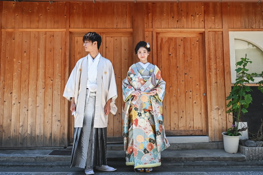 木造の格子壁の京町家を背に立つ、生成り色の着物袴の新郎と扇と鶴の柄が美しい花柄着物の花嫁