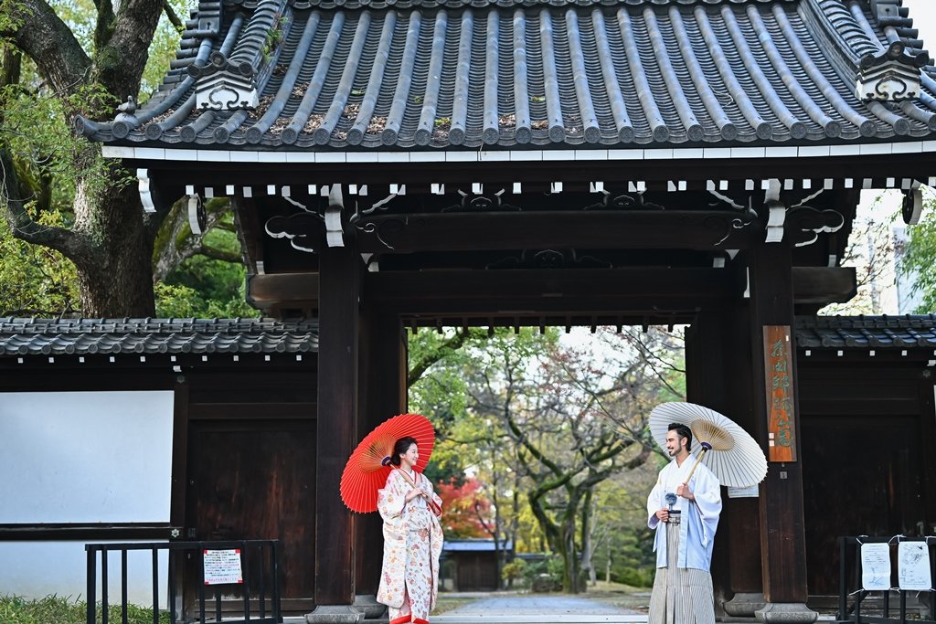 大きな瓦屋根の門の下、朱色の和傘をさす花嫁と白い和傘をさす新郎が向かい合って立つ