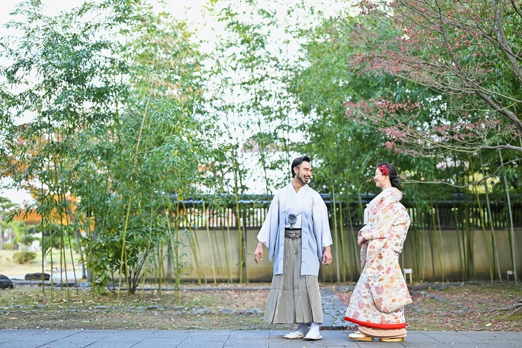 青々とした竹林を背景に、笑い合いながら向き合う水色の着物袴の新郎と生成り色の花柄着物の花嫁