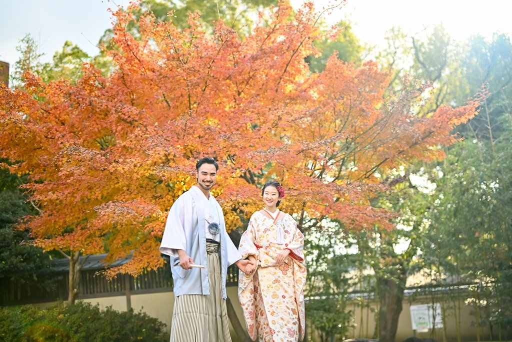 燃えるような紅葉の大木を背に手をつなぐ、灰色の着物袴の新郎と生成り色の花柄着物の花嫁