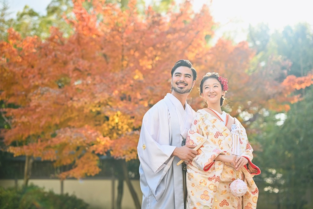 オレンジ色の紅葉を背に、遠くを見つめ微笑む灰色の着物袴の新郎と、髪に赤い花飾りとピンクの手鞠を付けた生成り色の着物姿の花嫁