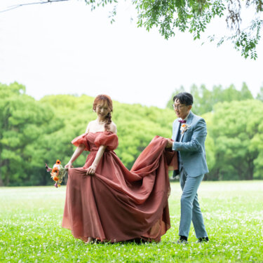 新緑の木陰の芝生で、テラコッタドレスの裾を新郎が持ち上げて一緒に歩く新郎新婦。公園でのロケーションフォト