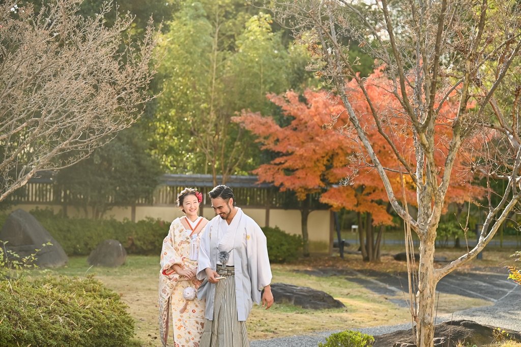 紅葉の木の下、庭園の小道を寄り添って歩く白い着物袴の新郎と生成り色の花柄着物の花嫁