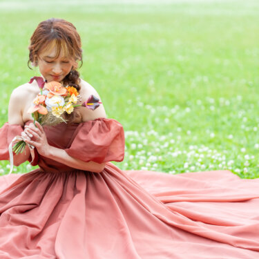 緑の芝生にドレスの裾を大きく広げて座り、オレンジのブーケに顔を寄せる花嫁。カラードレスの前撮り