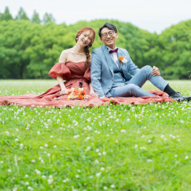 シロツメクサの咲く芝生に並んで座り、カメラに笑顔を向けるテラコッタドレスの花嫁とライトブルースーツの新郎。ガーデンフォトウェディング