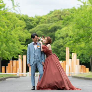新緑の並木道で、ブーケを新郎の頬に寄せてじゃれ合うテラコッタのカラードレスの花嫁とライトブルースーツの新郎。公園ロケーションフォト