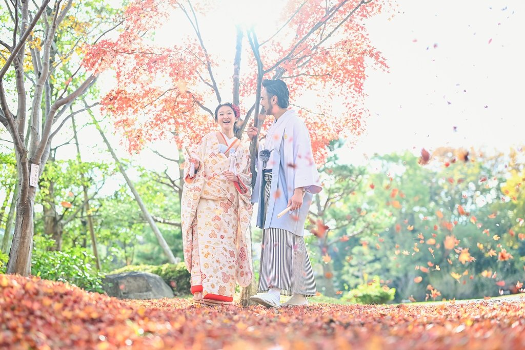 落ち葉が絨毯のように地面を覆う紅葉の中、舞い散る葉に囲まれ笑い合う生成り色の花柄着物の花嫁と灰色の着物袴の新郎