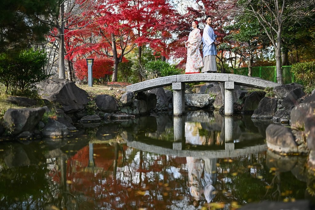 真っ赤な紅葉に囲まれた池の石橋の上に立つ、生成り色の着物姿の花嫁と灰色の着物袴の新郎と、水面に映るふたりの姿