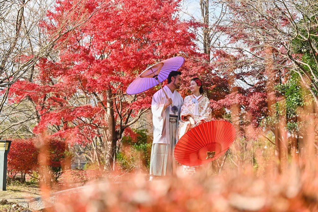 真っ赤に染まった紅葉の中、紫色の和傘をさす新郎と、朱色の和傘を手にした生成り色の着物姿の花嫁
