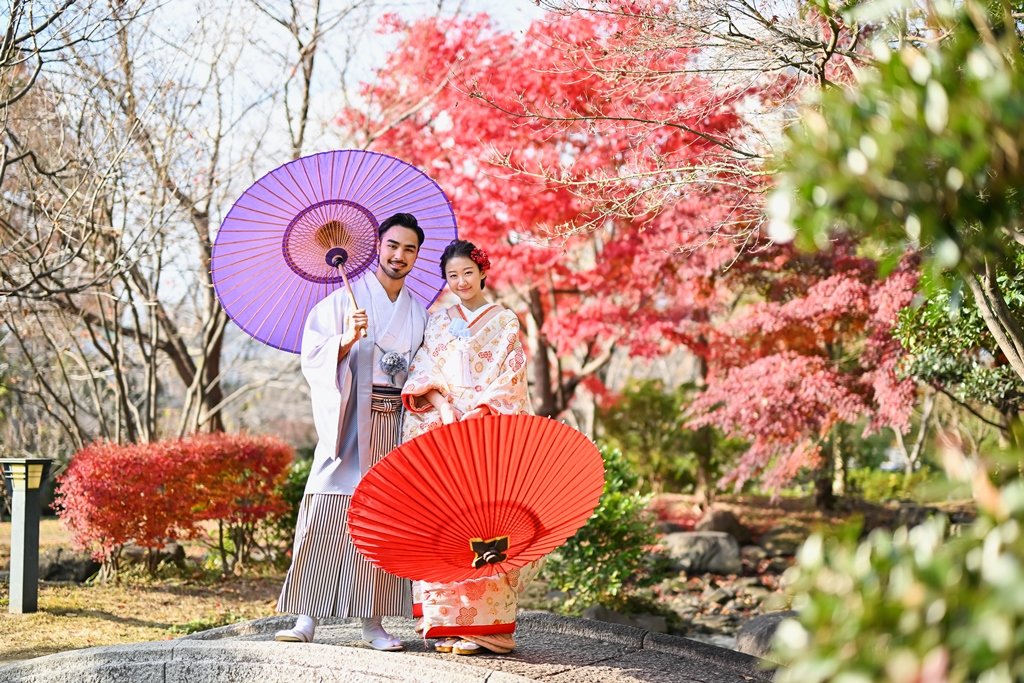 紅葉の庭園で寄り添い立つ、紫の和傘をさす灰色の着物袴の新郎と、朱色の和傘を持つ生成り色の花柄着物の花嫁