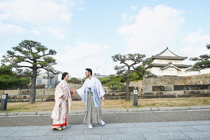 大阪城の櫓と石垣、松の木を背景に、石畳の上で手を取り合い向かい合う水色の着物袴の新郎と花柄着物の花嫁