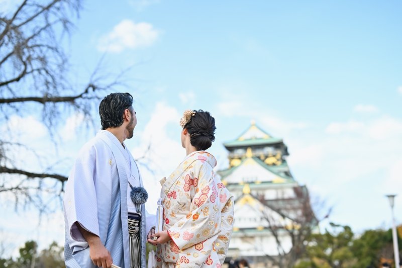 青空の下、大阪城を背景に見つめ合う水色の着物袴の新郎と、朱色の花柄模様が華やかな着物姿の花嫁