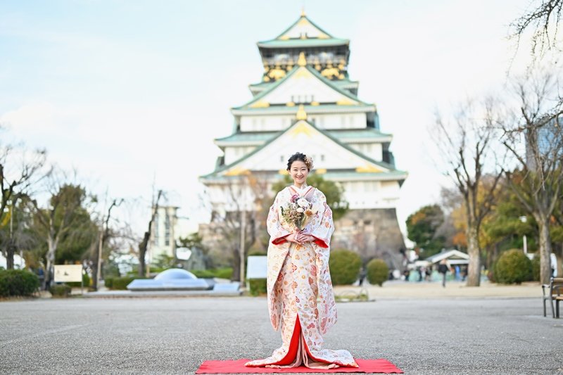 大阪城の天守閣を背に、赤い敷物の上に立つ花束を持った朱色の花柄着物姿の花嫁