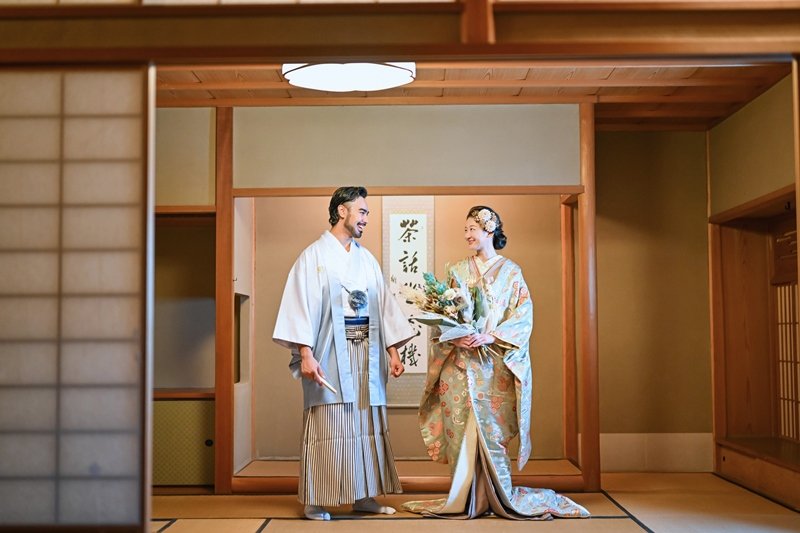 掛け軸のかかる畳の茶室で向き合う、白い羽織袴の新郎と、ブーケを手にした薄緑の花柄着物の花嫁