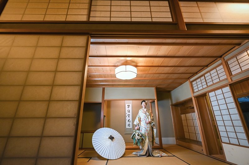 障子と畳の和室に佇む、ブーケを持った白と金の花柄着物姿の花嫁と、足元に置かれた白い和傘