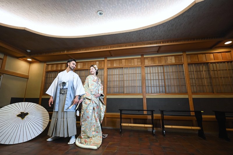 格子戸の広間で寄り添う、水色の紋付袴の新郎と金地に花柄の着物を着た花嫁、傍らに置かれた白い和傘