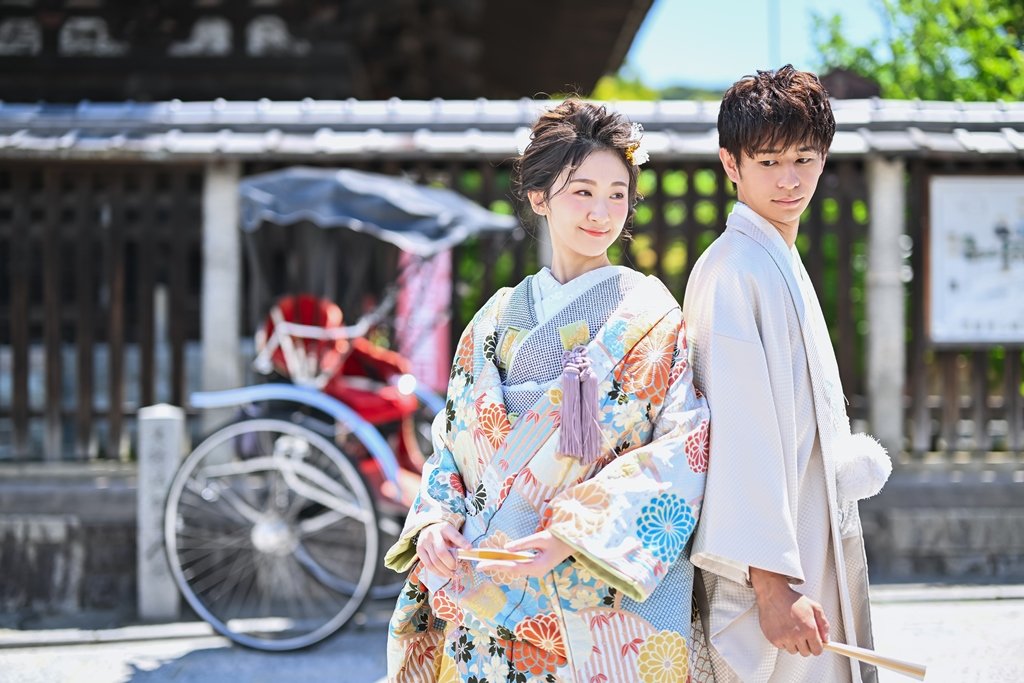 人力車を背景に背中合わせに立つ、生成り色の着物袴の新郎と菊柄が彩る花柄着物の花嫁