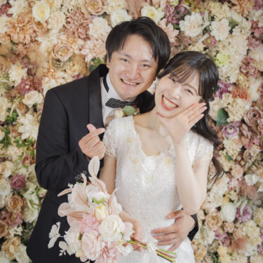 ピンクの花のフラワーウォールの前で頬に手を添えて笑うウェディングドレスの花嫁と黒いタキシードの新郎の縦位置の一枚。心斎橋店のスタジオで撮る洋装前撮り