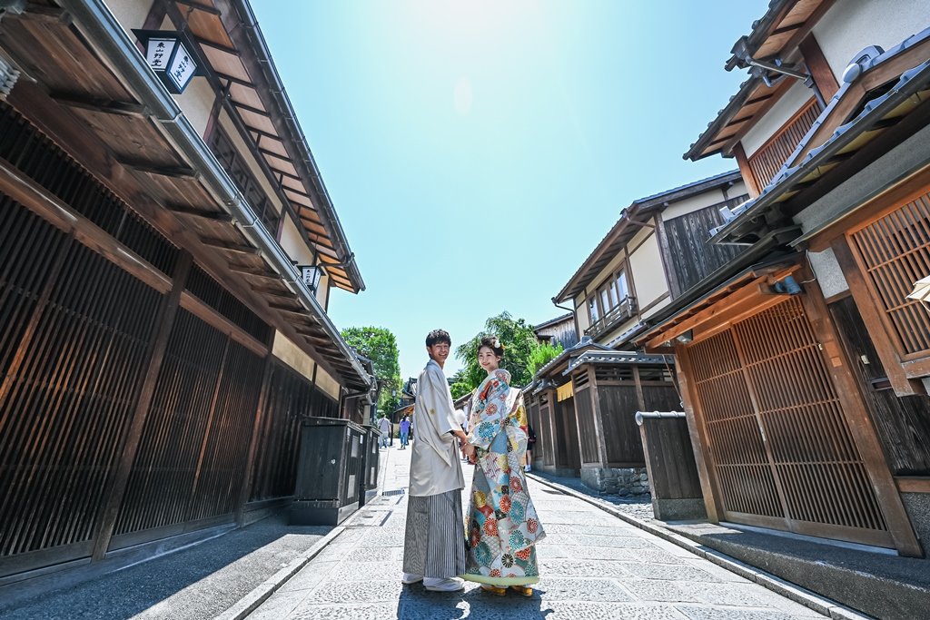 格子戸の京町家が両側に並ぶ石畳の通りの中央で、手をつなぎ振り返る生成り色の着物袴の新郎と花柄着物の花嫁