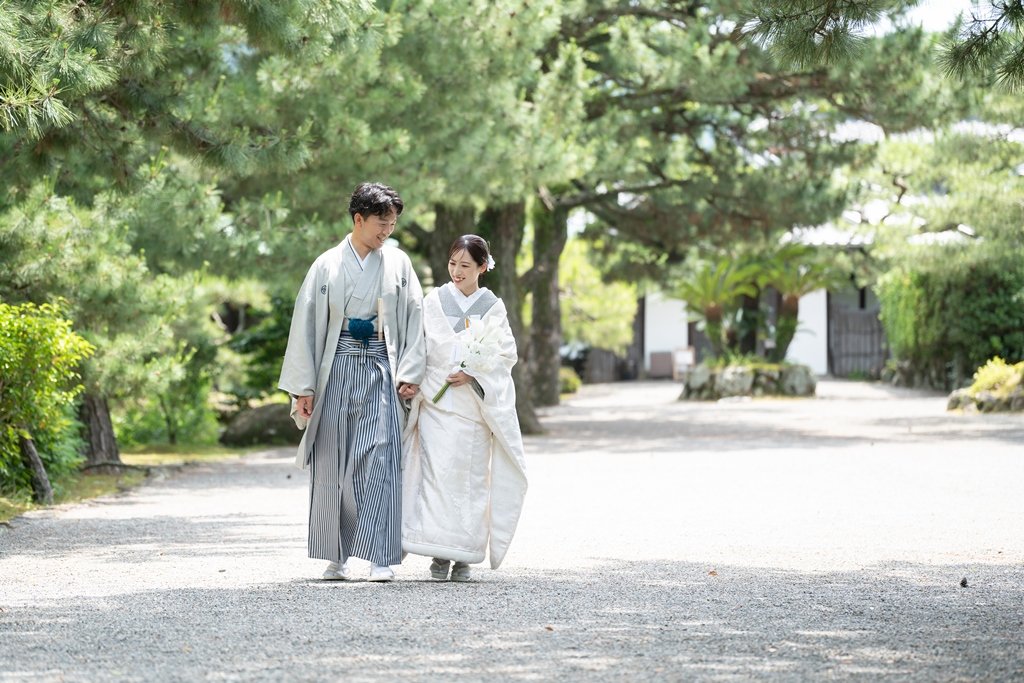 松の木々が並ぶ砂利道を、花束を手に手をつないで歩く生成り色の着物姿の花嫁と灰色の着物袴の新郎