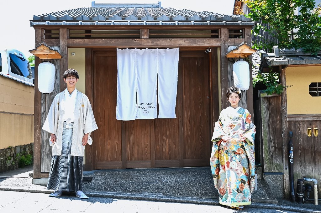 白い提灯が下がる京都の町家の店先で、笑顔で並んで立つ生成り色の着物袴の新郎と花柄着物の花嫁
