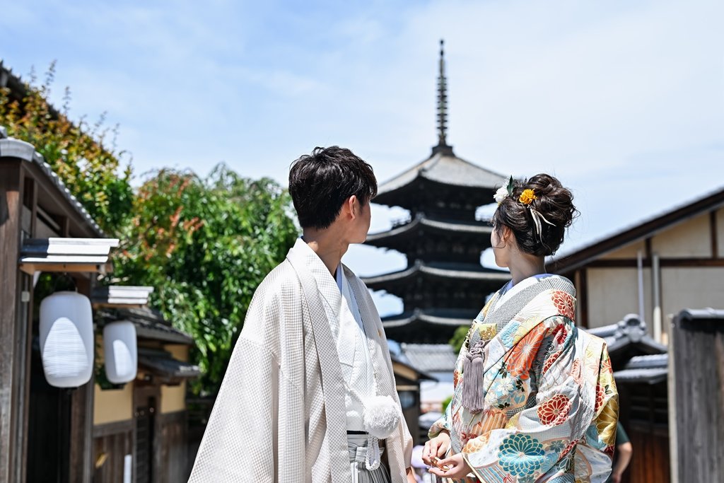 青空の下、白提灯が下がる京都の街並みと八坂の塔を背景に向かい合う生成り色の着物袴の新郎と花柄着物の花嫁