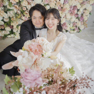 一面の花の壁を背に、ブーケを手にして寄り添い笑顔を見せる新郎新婦。フラワーウォールのスタジオフォトウェディング