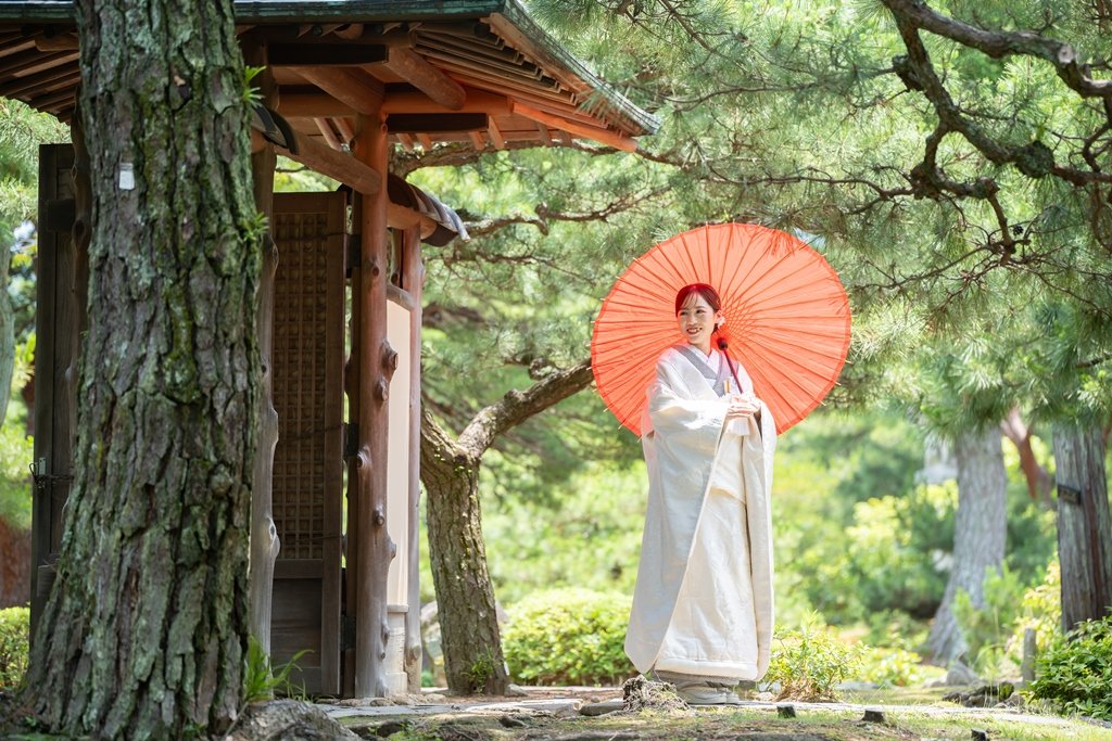 松の木々が茂る庭園の小さな門の前で、朱色の和傘をさして微笑む生成り色の着物姿の花嫁