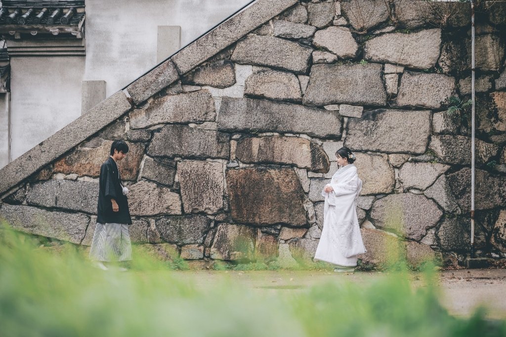 巨大な石垣を背に、黒紋付袴の新郎と白無垢を着た花嫁が向き合って立つ