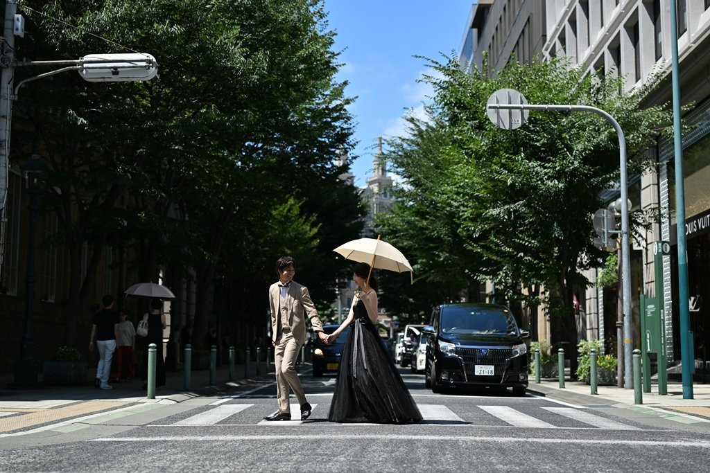 街路樹が並ぶ神戸旧居留地の横断歩道を手をつないで渡る、ベージュのスーツの新郎と黒いドレスにパラソルの花嫁
