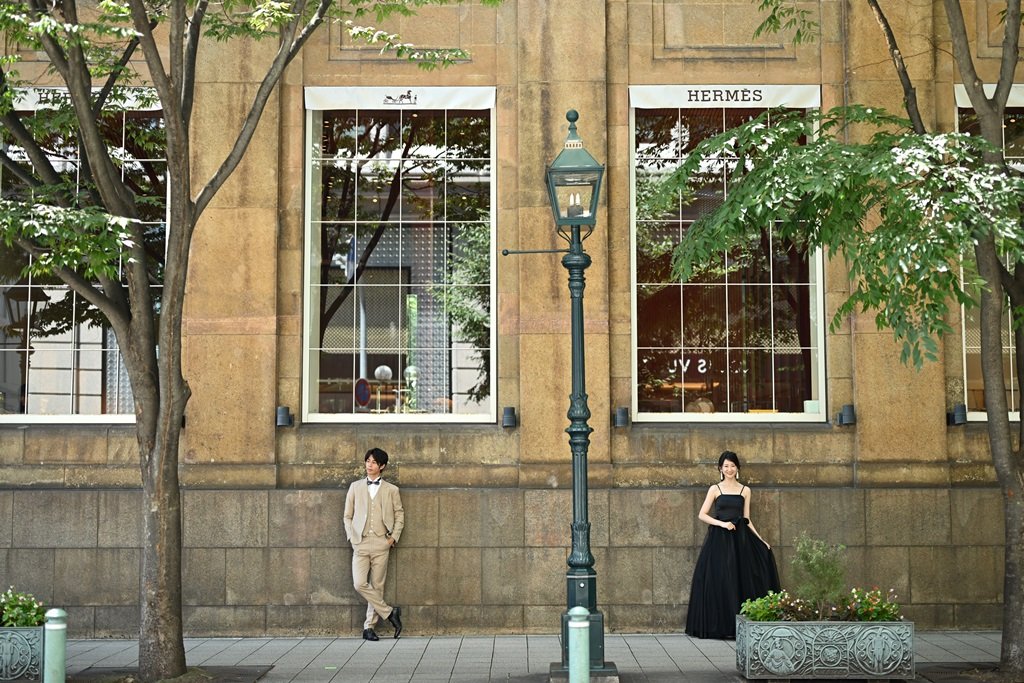 HERMÈSの店舗が並ぶ神戸旧居留地のガス灯の前で、離れて立つベージュのスーツの新郎と黒いドレスの花嫁