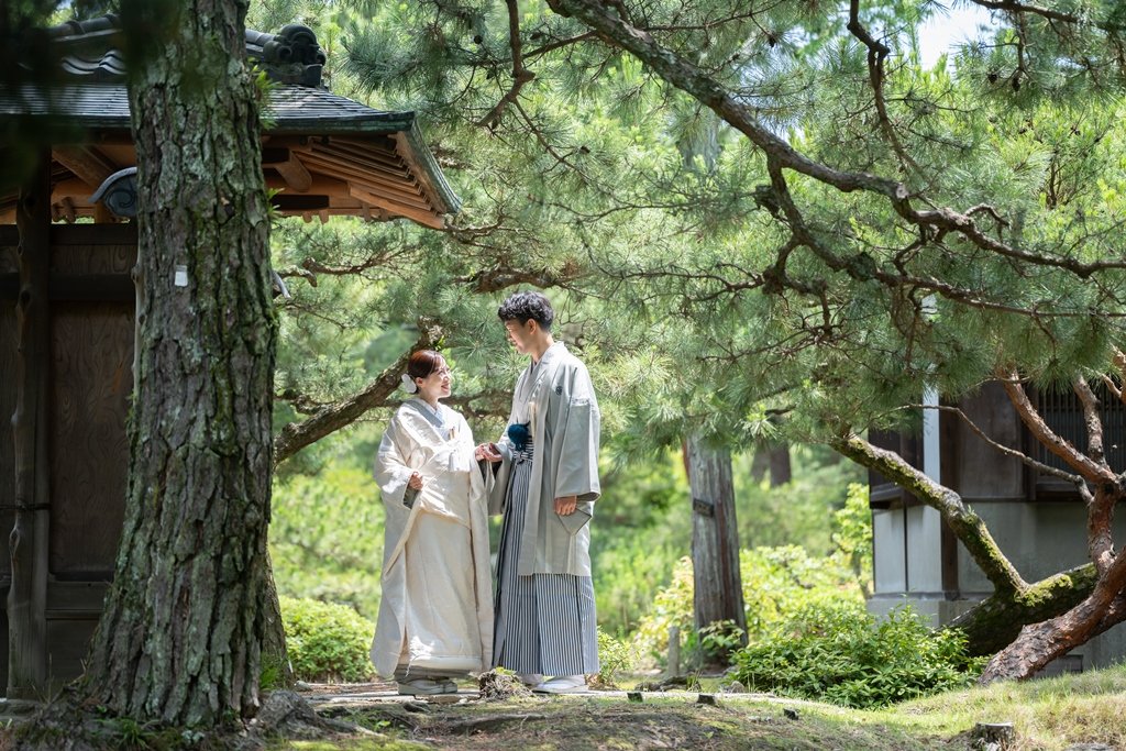 大きな松の枝が張り出す門の前で、手を取り合い見つめ合う灰色の着物袴の新郎と生成り色の着物姿の花嫁