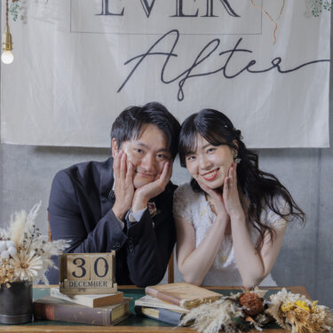 『Happily Ever After』のタペストリーとドライフラワーを飾ったテーブルで、両手で顔を包んで笑う黒いタキシードの新郎とレースのウェディングドレスの花嫁。日付ブロックを添えた洋装前撮り