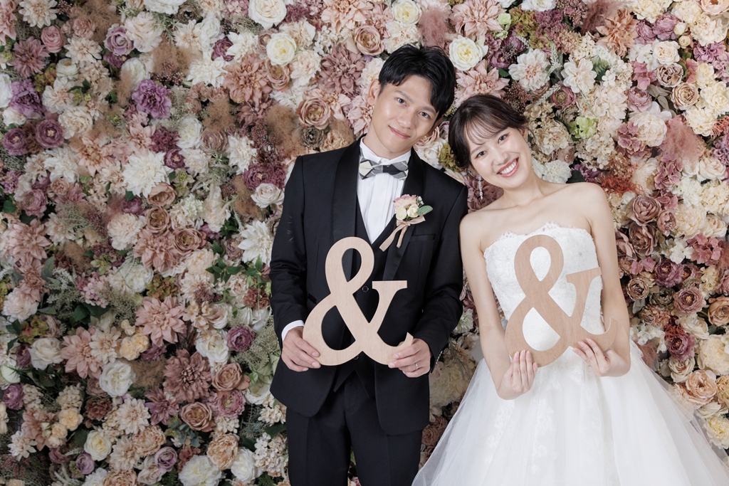 花の壁を背景に木製の「&」オブジェを持つ新郎新婦