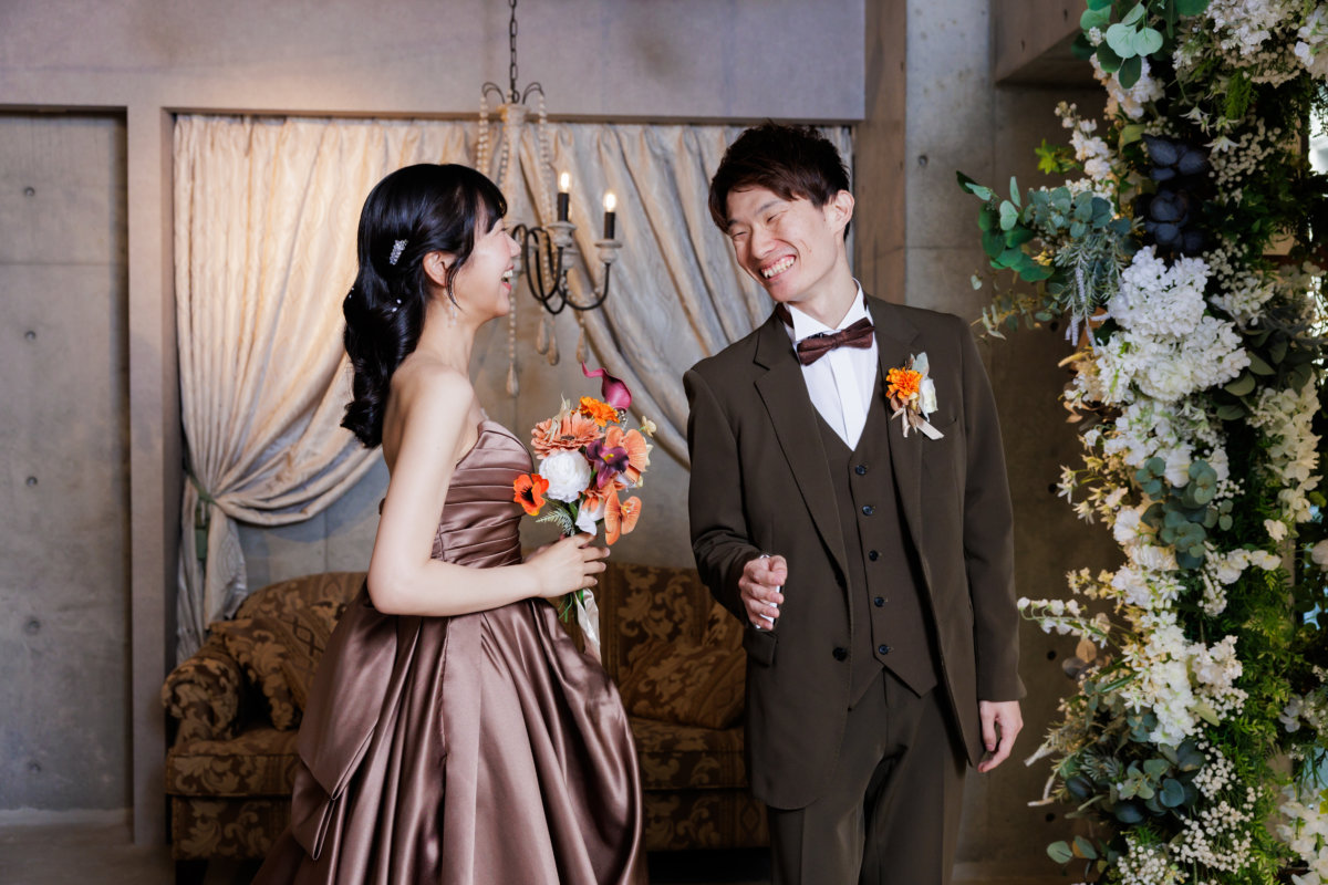 白いカーテンとシャンデリアのスタジオで、ブーケを持って向かい合うブラウンのカラードレスの花嫁とブラウンのスーツの新郎。心斎橋店のスタジオで撮る洋装前撮り
