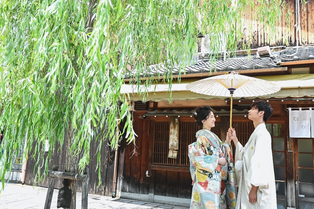 しだれ柳の下、白い和傘を差しかけながら見つめ合う生成り色の着物袴の新郎と花柄着物の花嫁
