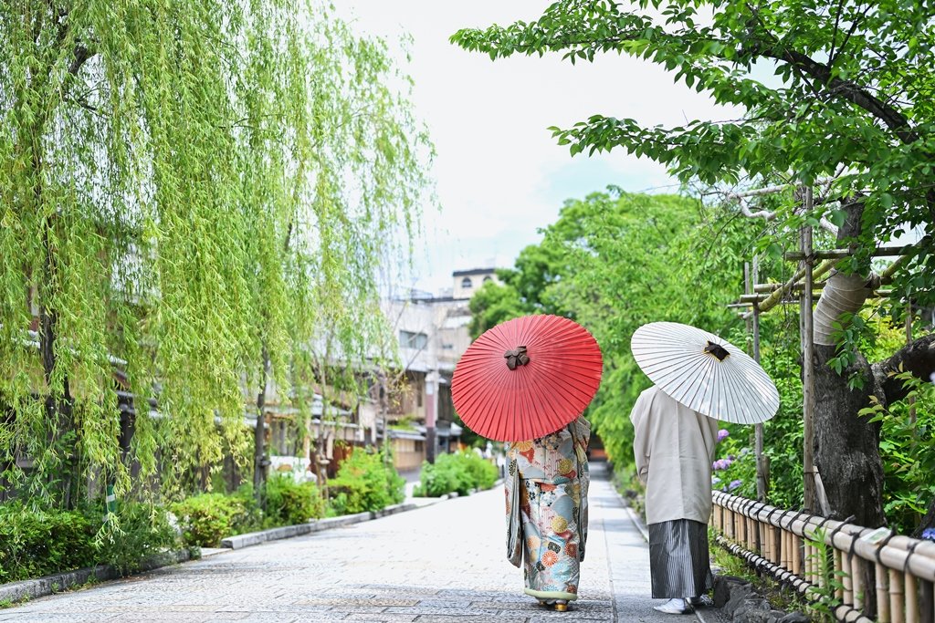 しだれ柳が揺れる祇園の石畳の道を、朱色と白の和傘をさして歩く花柄着物の花嫁と生成り色の着物袴の新郎の後ろ姿