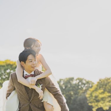 新郎が花嫁をおんぶして笑い合うワンシーン。ブラウンのツイードスーツと白いウェディングドレスで撮る自然体な前撮り
