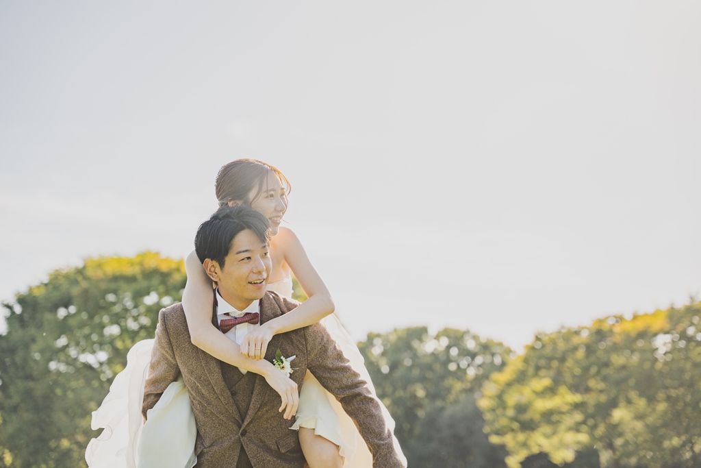 新郎が花嫁をおんぶして笑い合うワンシーン。ブラウンのツイードスーツと白いウェディングドレスで撮る自然体な前撮り