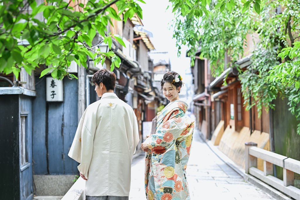 新緑の枝葉が茂る祇園の路地で、後ろを振り返り微笑む花柄着物の花嫁と、隣で佇む生成り色の着物袴の新郎