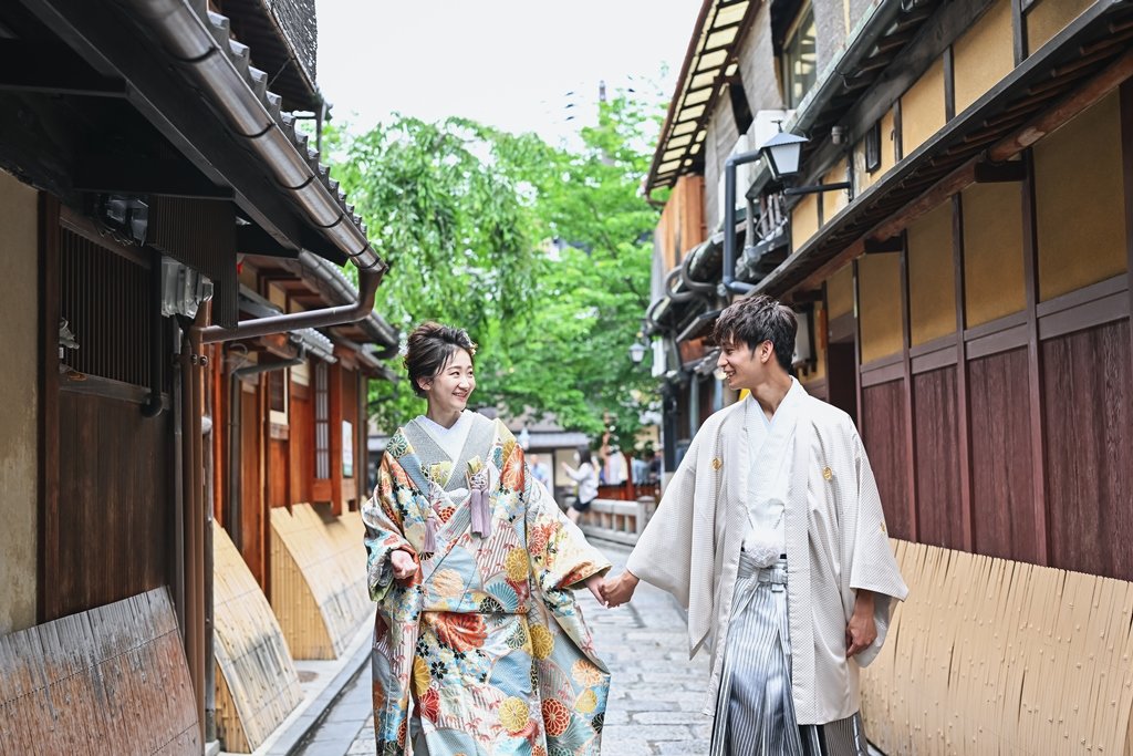 簾がかかる祇園の石畳の路地を、笑顔で手をつなぎ歩く花柄着物の花嫁と生成り色の着物袴の新郎