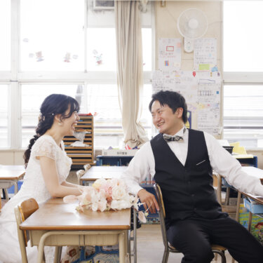 教室の机に着き、ブーケを置いて笑い合う白いウェディングドレスの花嫁と新郎。学校ロケーションフォト