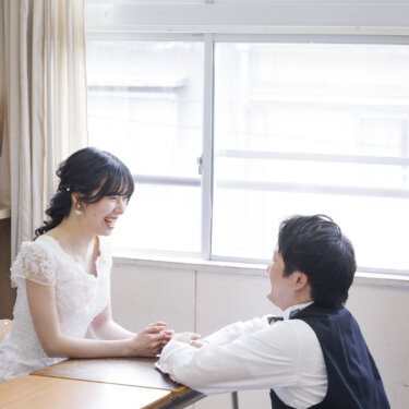教室の窓辺で机を挟んで向かい合い、手を取り合って笑い合う白いウェディングドレスの花嫁とベスト姿の新郎。学校ロケのフォトウェディング