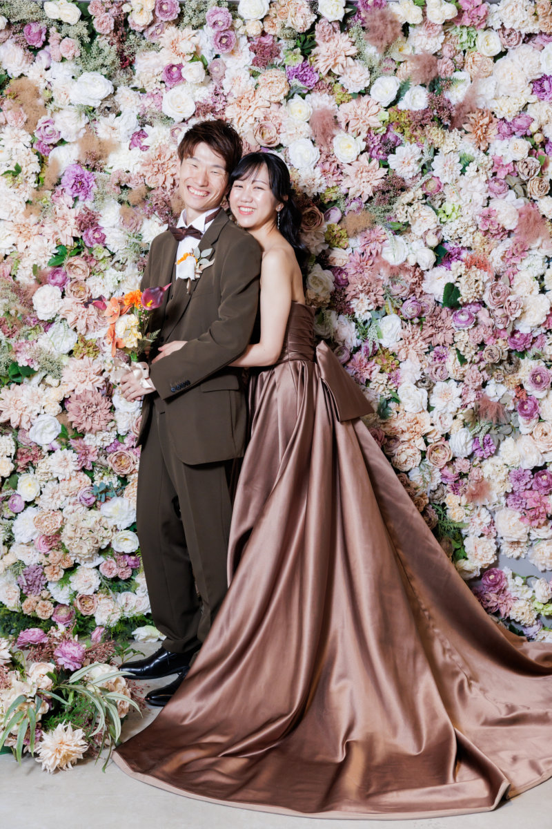 ピンクとベージュの花で埋め尽くしたフラワーウォールの前で寄り添うブラウンのスーツの新郎とブラウンのカラードレスの花嫁。心斎橋店のスタジオで撮る洋装前撮り