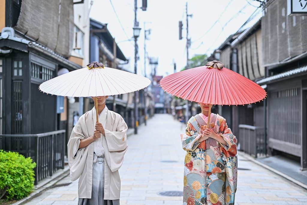 白い和傘と朱桃色の和傘で顔を隠すように立つ、生成り色の着物袴の新郎と花柄着物の花嫁、祇園の路地を背景にしたアートなショット