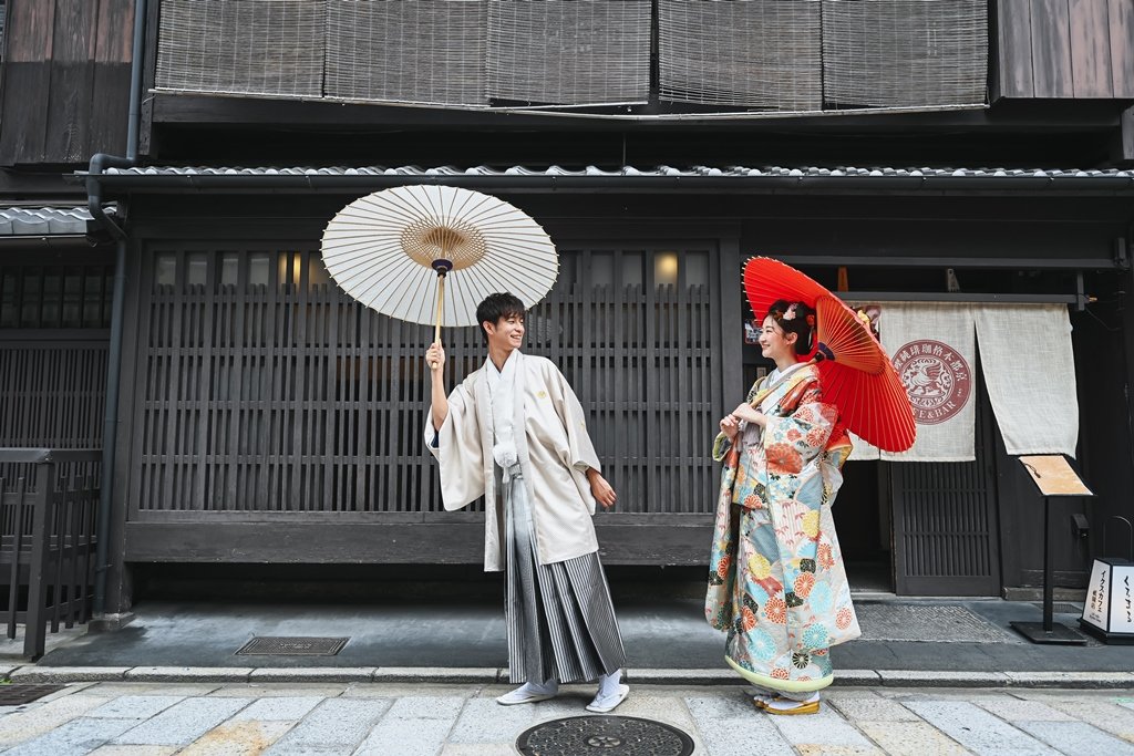簾が下がる祇園の趣ある町家を背に、白い和傘をさす生成り色の着物袴の新郎と朱色の和傘をさす花柄着物の花嫁