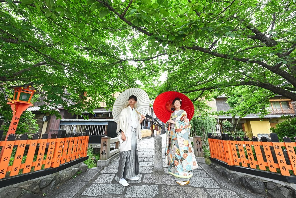 新緑のトンネルの下、祇園巽橋のたもとで白い和傘をさす生成り色の着物袴の新郎と朱色の和傘をさす花柄着物の花嫁
